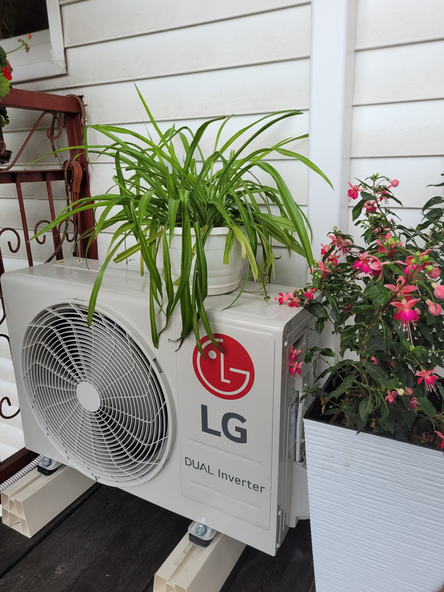 Zewnętrzna jednostka klimatyzacji LG Dual Inverter ustawiona na plastikowych wspornikach, z doniczką z zielonymi roślinami na górze i fuksją w białej donicy obok, na tle białej drewnianej ściany.