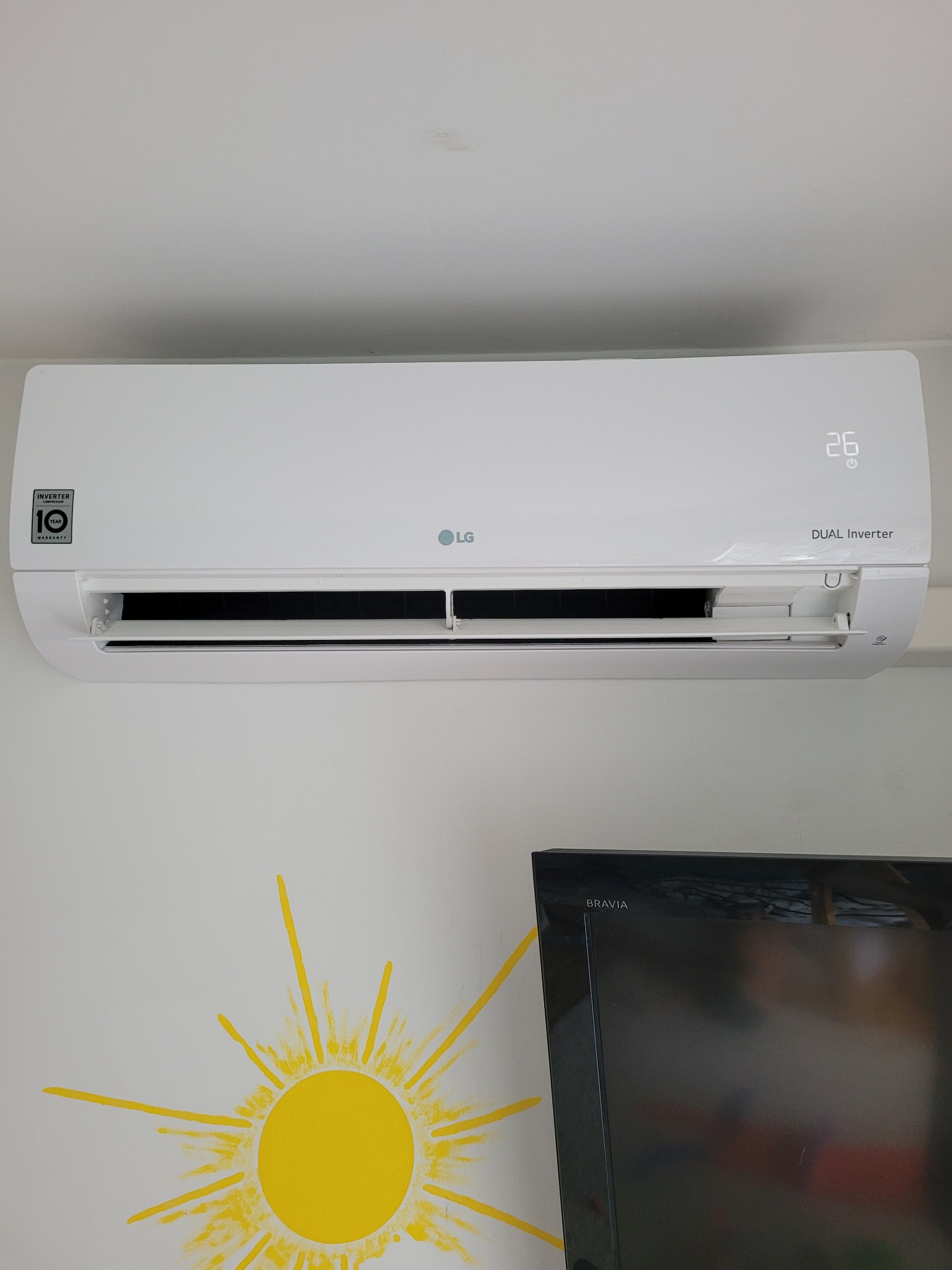 Zamontowana na ścianie biała klimatyzacja LG Dual Inverter z wyświetlaczem wskazującym 26 stopni, poniżej widoczny fragment telewizora Bravia i żółta dekoracja ścienna w kształcie słońca.