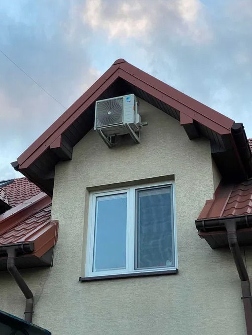 Zewnętrzna jednostka klimatyzacji zamontowana na ścianie szczytowej domu, widoczna pod okapem dachu, z oknem poniżej. Na ścianie widoczna faktura tynku.
