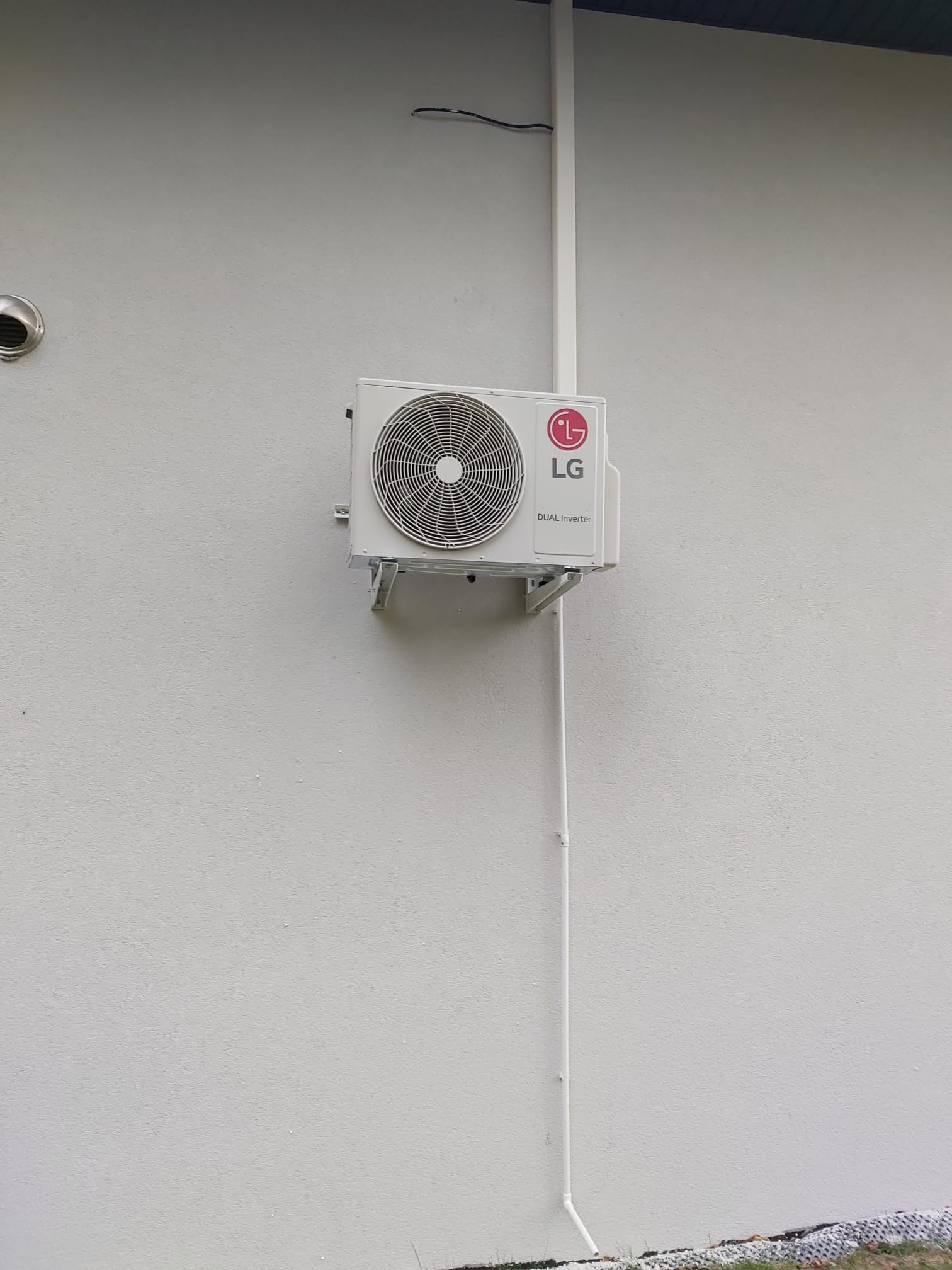 Biała jednostka zewnętrzna klimatyzacji LG Dual Inverter zamontowana na szarej, teksturowanej ścianie budynku, z widocznymi przewodami w białej osłonie.