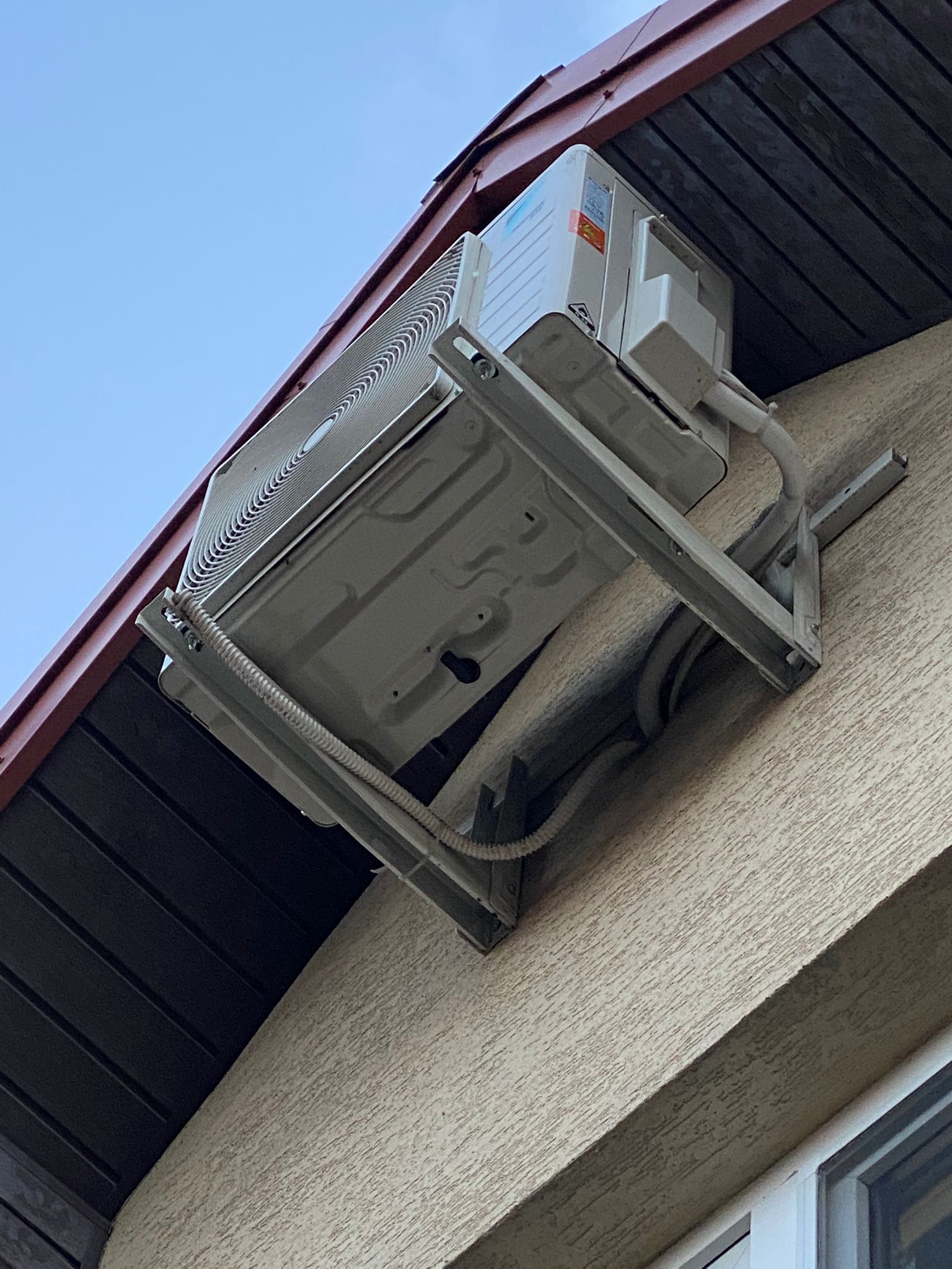 Zewnętrzna jednostka klimatyzacji zamontowana na wsporniku na elewacji budynku pod okapem dachu.