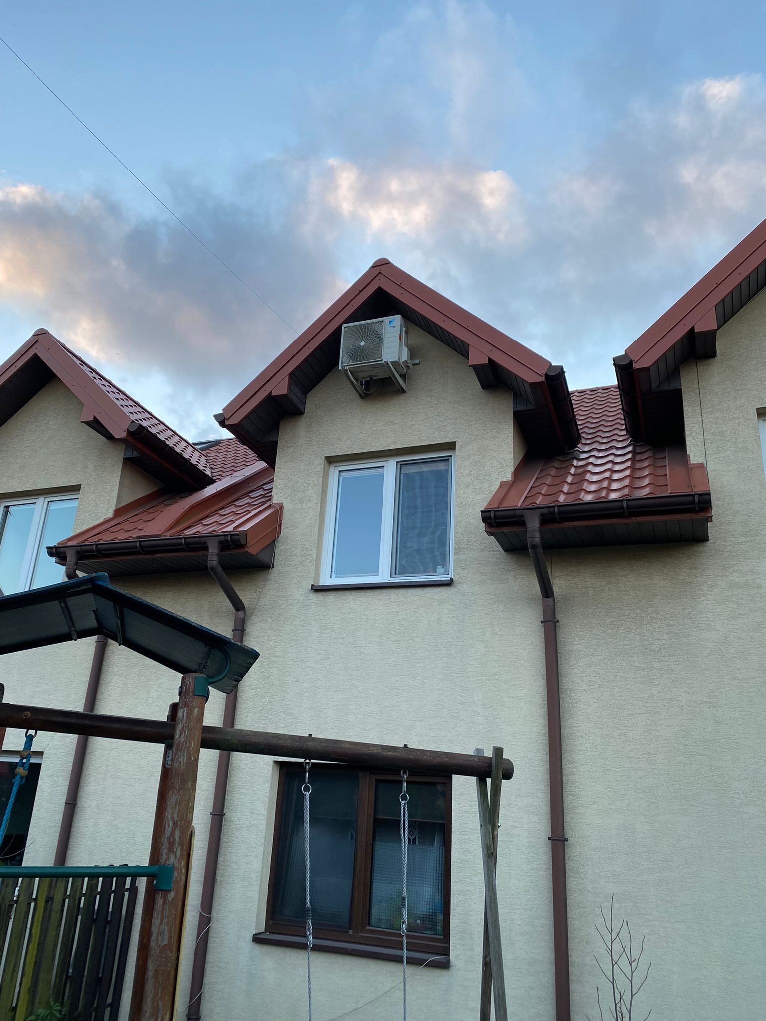 Elewacja domu z zamontowanym klimatyzatorem na poddaszu, widoczne brązowe rynny i fragment huśtawki.