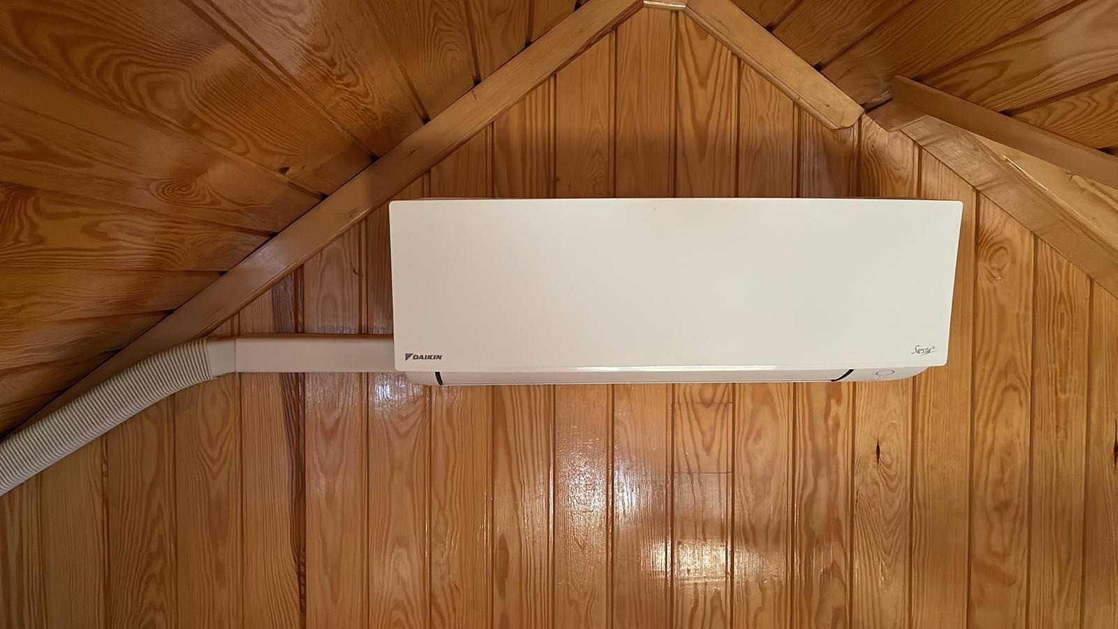 Biały klimatyzator Daikin Siesta zamontowany na drewnianej ścianie z boazerii i drewnianym sufitem pod skosem.