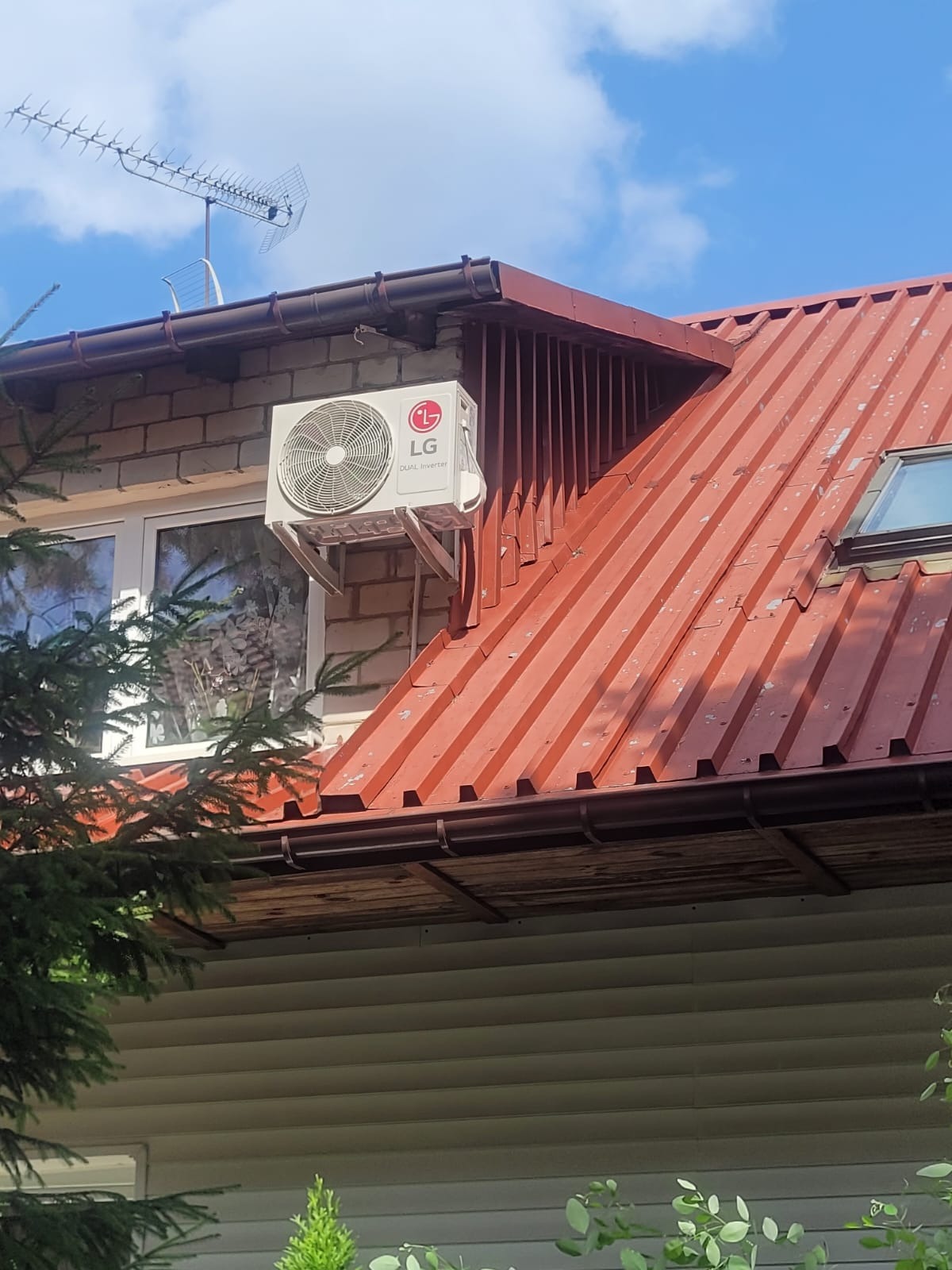 Zewnętrzna jednostka klimatyzacji LG przymocowana do ściany budynku z cegły, widoczna antena telewizyjna na dachu.