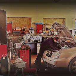 Mechanik pochylony nad silnikiem otwartego samochodu w warsztacie, widoczne narzędzia i wyposażenie warsztatowe w tle.