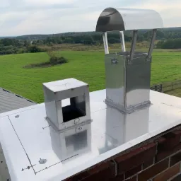 Nowe, błyszczące, metalowe zakończenie komina z daszkiem ochronnym, zamontowane na ceglanym kominie, z widocznym krajobrazem pól i lasów w tle.