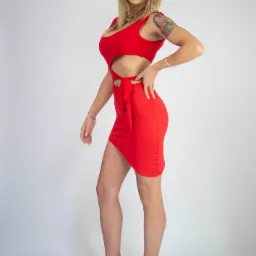 Kobieta w czerwonej, dopasowanej sukience z wycięciami, pozująca z ręką na biodrze, widoczny tatuaż na ramieniu i beżowe buty na platformie.