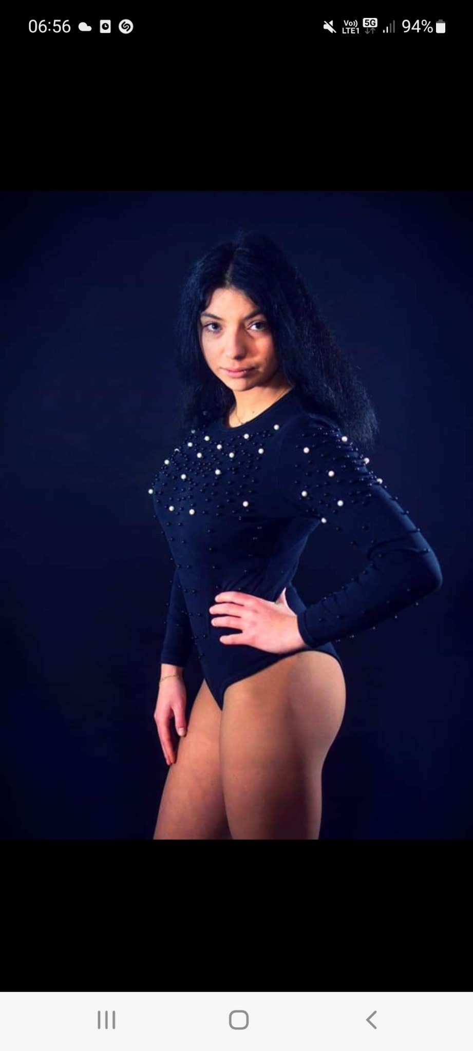 Portret kobiety w czarnym body zdobionym perłami, pozującej na ciemnym tle z ręką na biodrze.