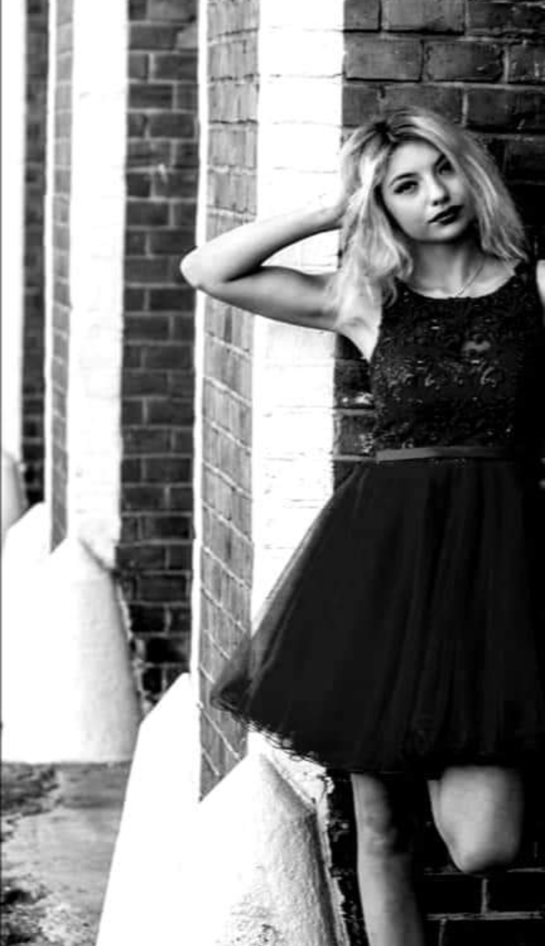 Czarno-białe zdjęcie młodej kobiety w ozdobnej sukience pozującej przy ceglanym murze, z ręką uniesioną do włosów.