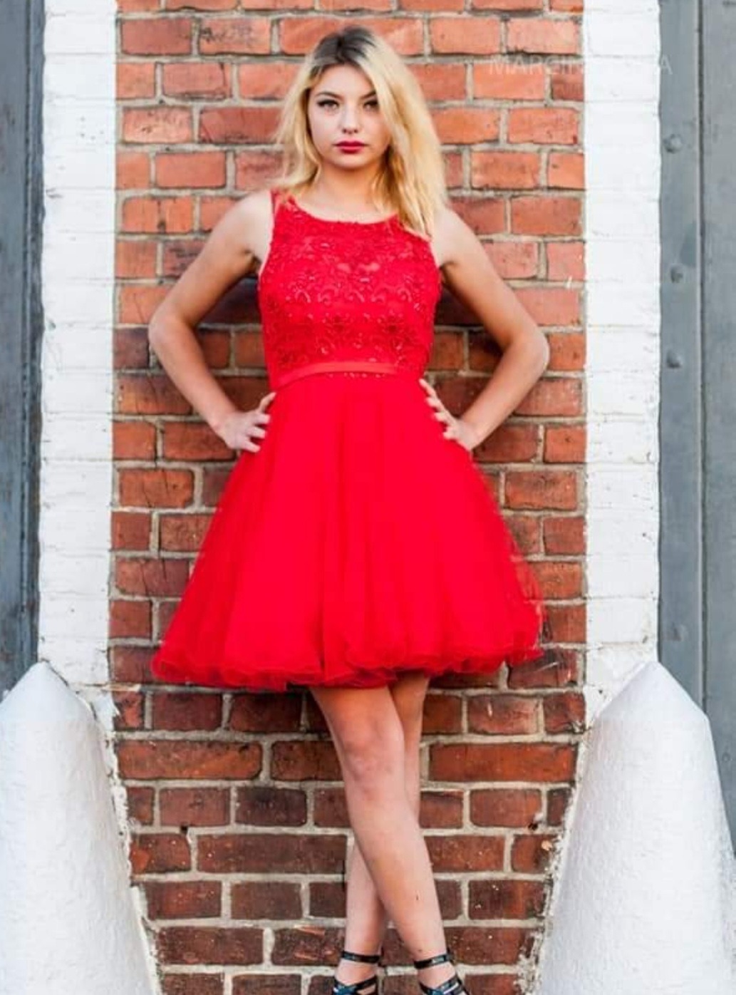 Młoda blondynka w czerwonej sukience z koronkową górą pozuje na tle ceglanej ściany z białymi elementami. Uśmiech i pewna postawa modelki.