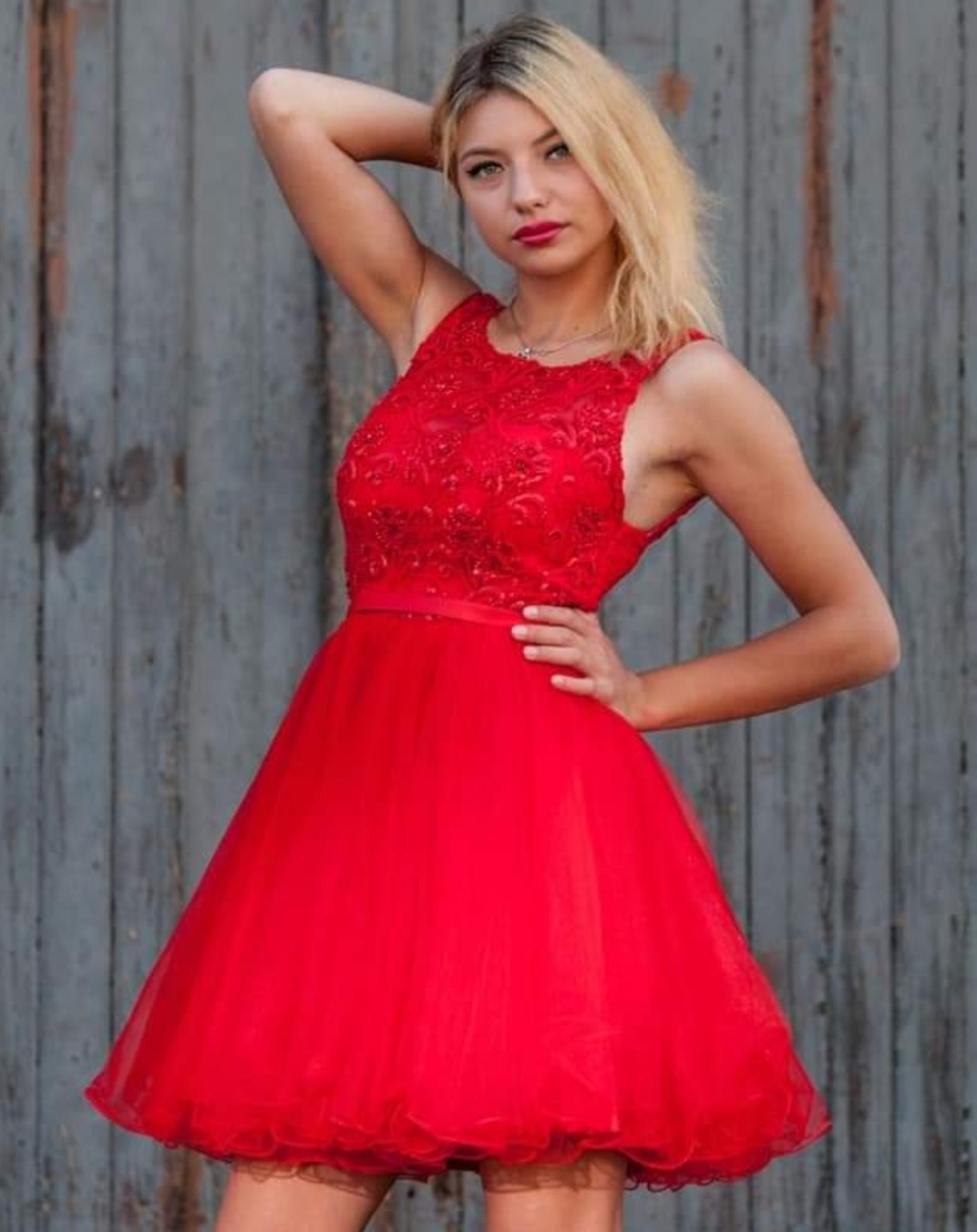 Portret młodej kobiety w czerwonej, balowej sukience z koronkową górą, pozującej na tle szarej, drewnianej ściany.