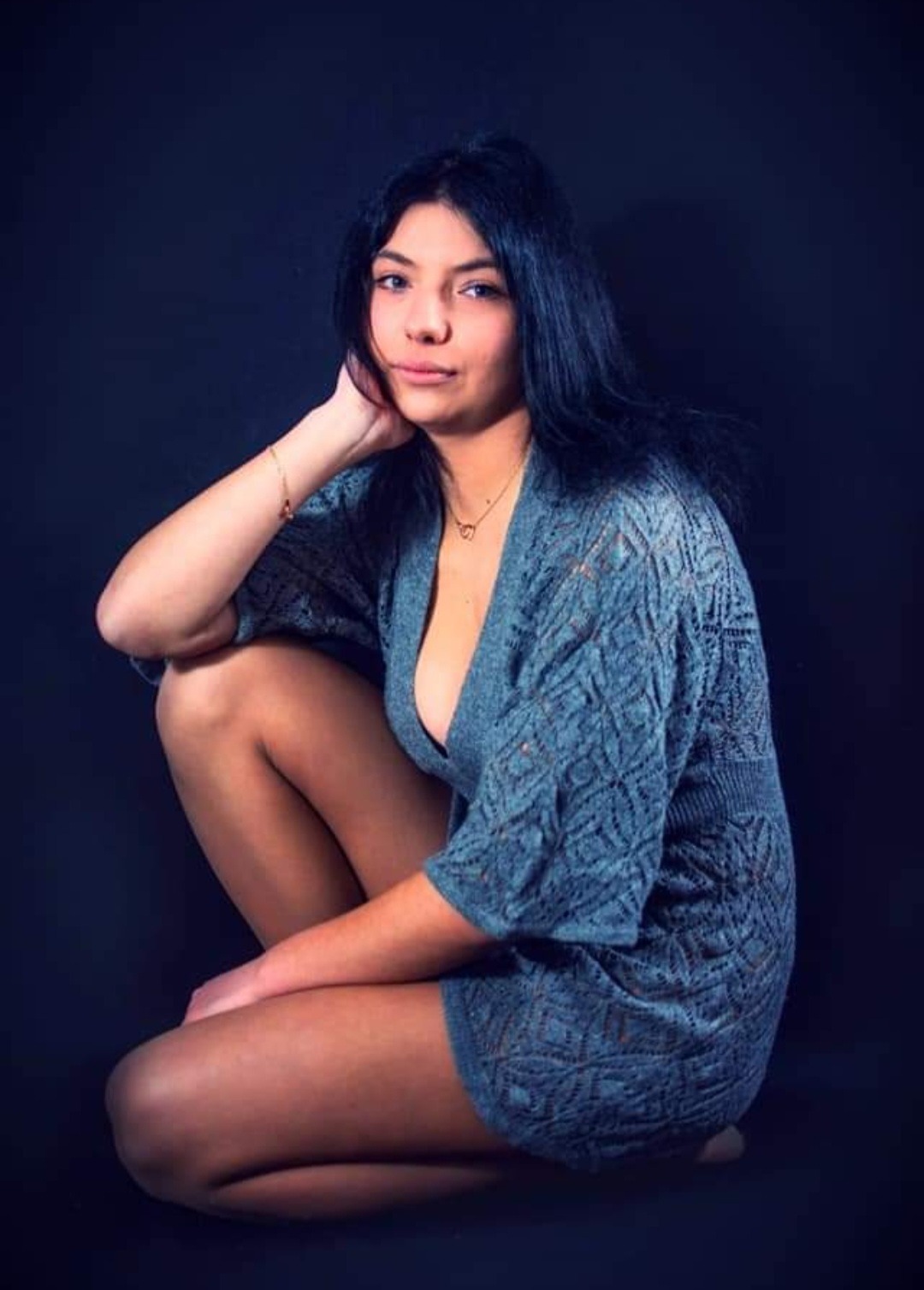 Portret kobiety w pozycji siedzącej, ubrana w ażurowy sweter w odcieniach szarości, z delikatną biżuterią i ciemnym tłem.