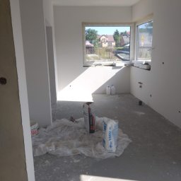 Rafcio - Surowe wnętrze w trakcie prac wykończeniowych, widoczne worki z zaprawą, folia ochronna na podłodze, świeżo pomalowane ściany i widok na zewnątrz przez okno.