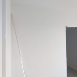 Rafcio - Świeżo pomalowana, biała ściana w rogu pomieszczenia, widoczna listwa przypodłogowa i fragment otworu drzwiowego z szarym wnętrzem w tle.