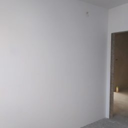 Rafcio - Świeżo pomalowana, gładka, biała ściana w rogu pomieszczenia, widoczny surowy betonowy oścież drzwiowy i fragment podłogi z resztkami po remoncie.