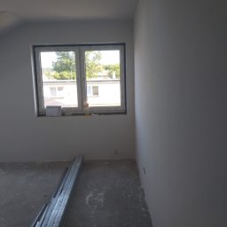 Rafcio - Wnętrze pokoju po szpachlowaniu ścian na gładko, z widocznymi profilami do płyt karton-gips na podłodze i oknem z widokiem na budynek mieszkalny.