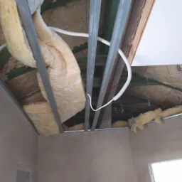 Montaż izolacji z wełny mineralnej w konstrukcji metalowej podwieszanego sufitu, widoczne kable elektryczne i nieotynkowane ściany.