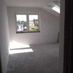 Pokój na poddaszu w trakcie remontu z widokiem na ogród przez okno, ściany świeżo pomalowane na biało, surowa podłoga przygotowana do dalszych prac wykończeniowych, widoczne ślady po szpachlowaniu.