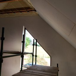 Rafcio - Poddasze w trakcie prac wykończeniowych: widoczne rusztowanie, okno o nietypowym kształcie, izolacja stropu i wstępnie wykończone ściany ze śladami szpachlowania.