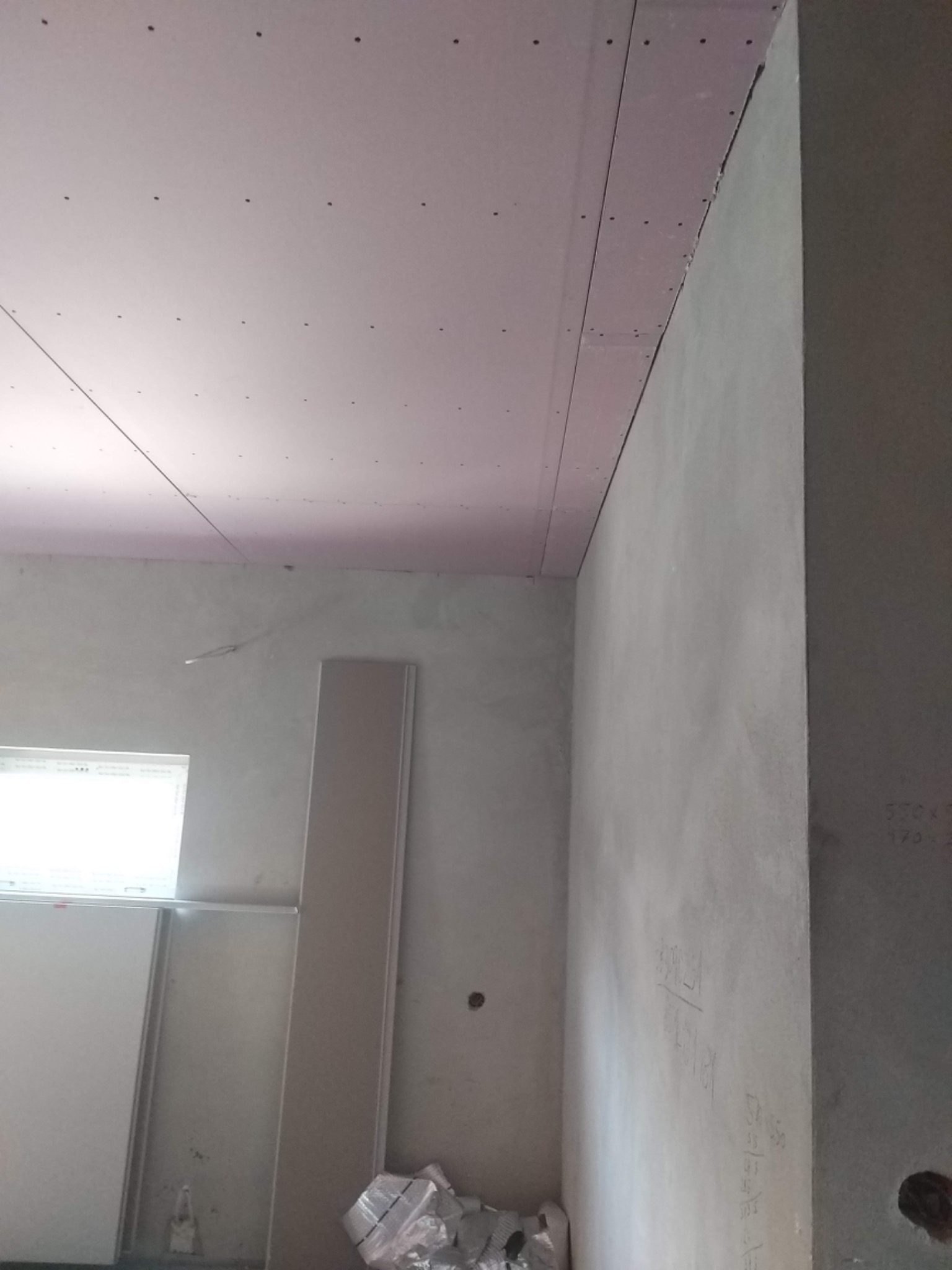 Wnętrze w trakcie prac wykończeniowych: zamontowany sufit podwieszany z płyt kartonowo-gipsowych, surowe ściany z widocznymi notatkami, materiały budowlane u dołu kadru.