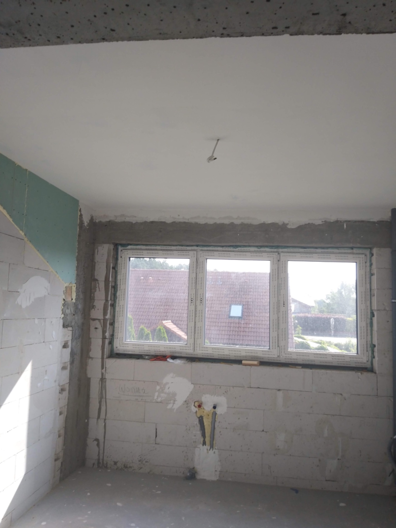 Surowy stan wnętrza w trakcie prac wykończeniowych: ściany z bloczków, częściowo zamontowana płyta na ścianie, okno z widokiem na zewnątrz, instalacja elektryczna wystająca ze ściany, surowy...