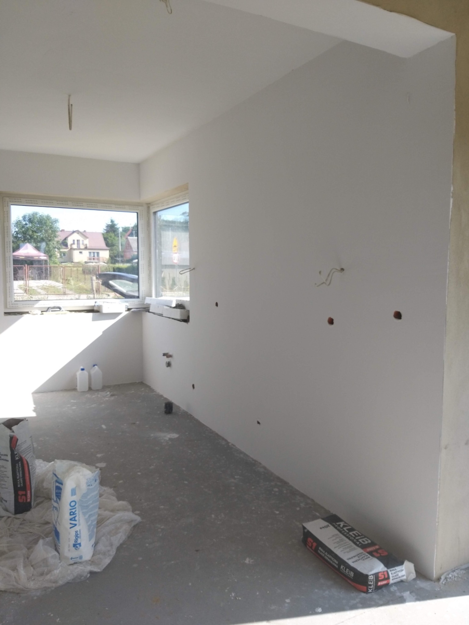Wnętrze w trakcie prac wykończeniowych: gładkie, pomalowane na biało ściany, widoczne puszki elektryczne, worki z gipsem i materiałami budowlanymi na podłodze, duże okno z widokiem na domy.