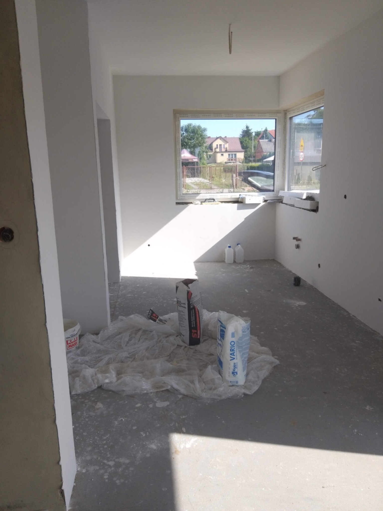 Surowe wnętrze w trakcie prac wykończeniowych, widoczne worki z zaprawą, folia ochronna na podłodze, świeżo pomalowane ściany i widok na zewnątrz przez okno.