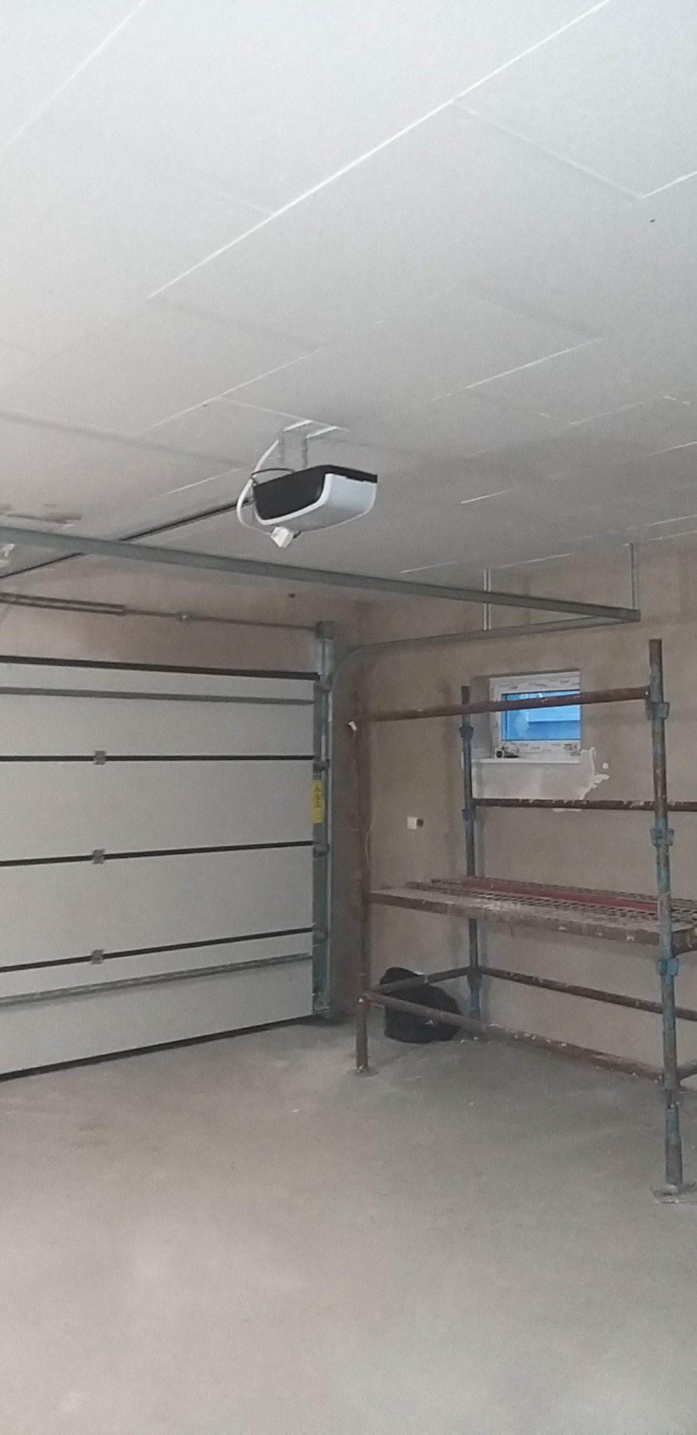 Wnętrze garażu w trakcie prac wykończeniowych: widoczne rusztowanie, brama garażowa segmentowa, zamontowany automat do bramy pod sufitem oraz sufit podwieszany z płyt kartonowo-gipsowych.