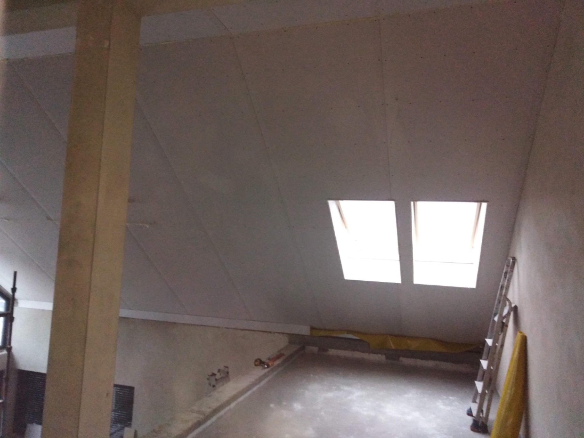 Poddasze w trakcie prac wykończeniowych: zamontowany sufit z płyt, widoczne okna dachowe, drabina oparta o ścianę i zabezpieczona folia malarska. W tle kolumna konstrukcyjna.