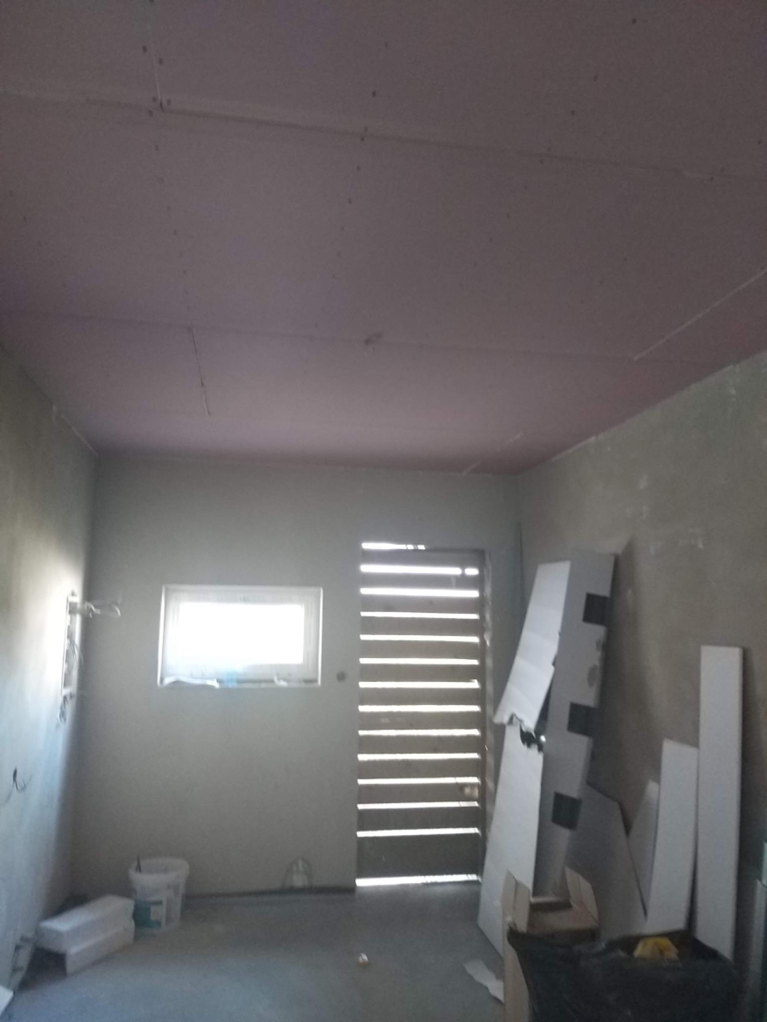 Pokój w trakcie prac wykończeniowych: surowa ściana, okno z widokiem na zewnątrz, zamontowany sufit z płyt, widoczne płyty kartonowo-gipsowe i wiadro na podłodze.