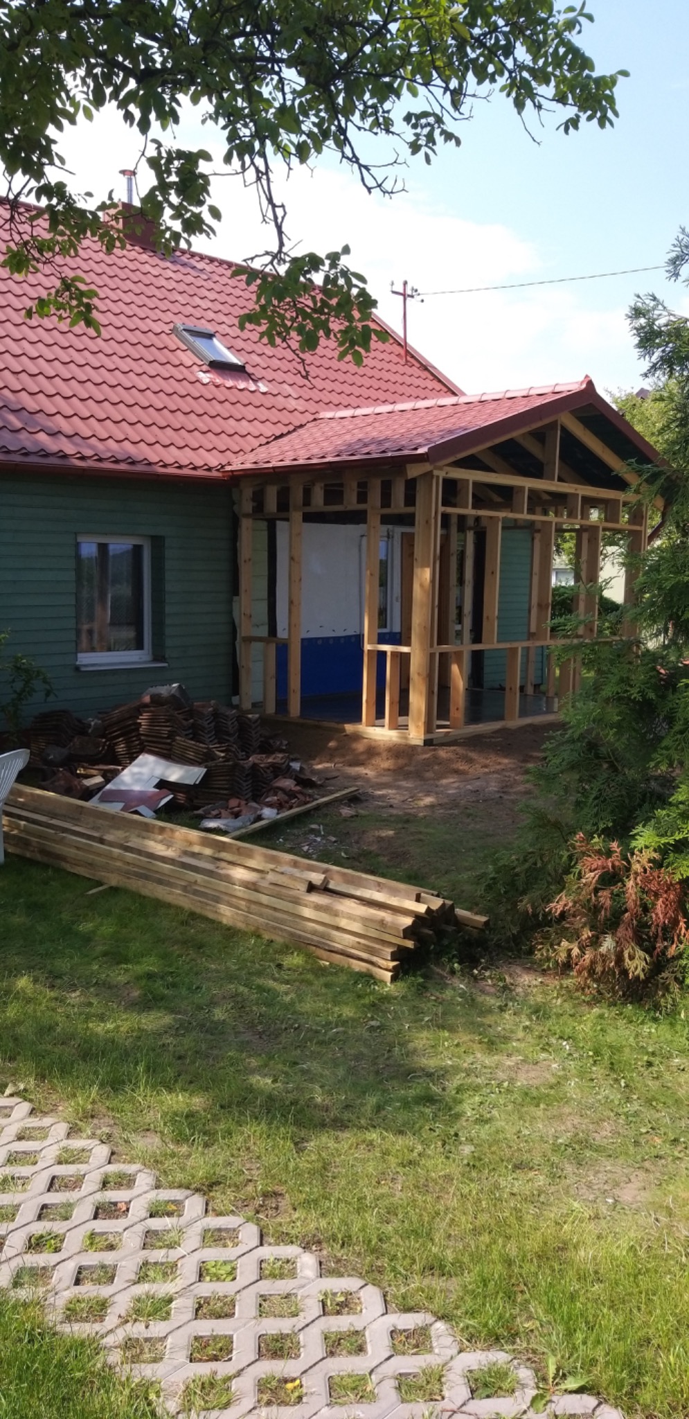 Budowa drewnianej konstrukcji werandy przy zielonym domu z czerwoną dachówką; widoczne materiały budowlane i fragment chodnika z ażurowych płyt.