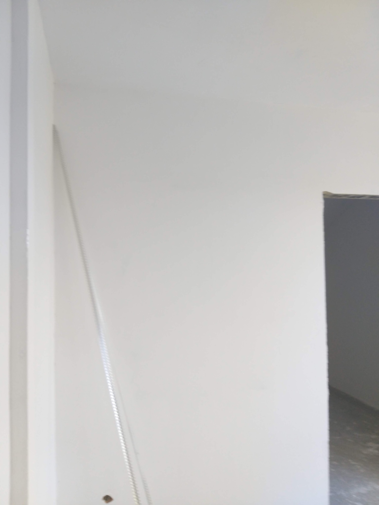 Świeżo pomalowana, biała ściana w rogu pomieszczenia, widoczna listwa przypodłogowa i fragment otworu drzwiowego z szarym wnętrzem w tle.