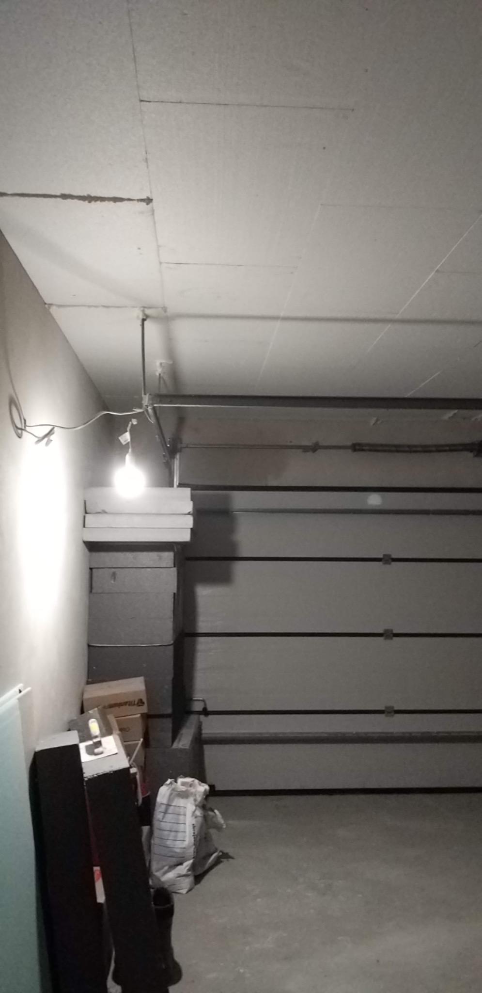 Niedokończony sufit z płyt kartonowo-gipsowych w garażu, widoczne szpary między płytami, stos płyt na podłodze pod żarówką, brama garażowa w tle.