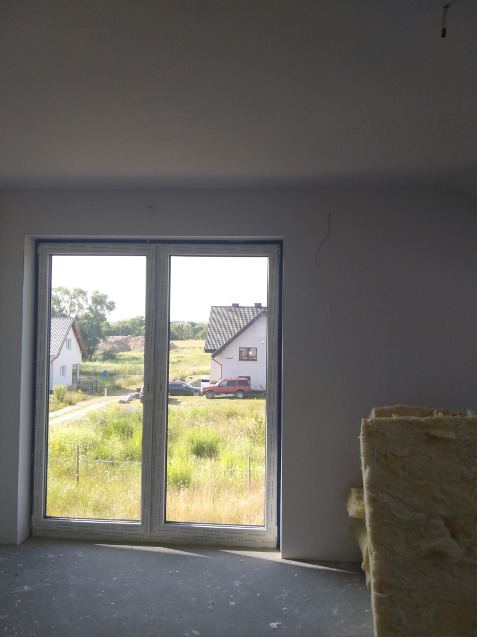 Surowe wnętrze z niezabezpieczonym oknem PCV, widok na zewnątrz z dwoma domami i samochodami, przy ścianie leżą płyty izolacyjne, na ścianie widoczne ślady po instalacjach.