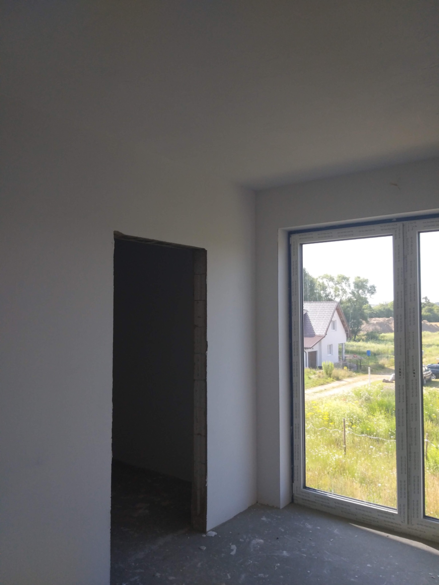 Surowe wnętrze w trakcie prac wykończeniowych: białe ściany, otwór drzwiowy bez ościeżnicy i duże okno z widokiem na dom i łąkę.