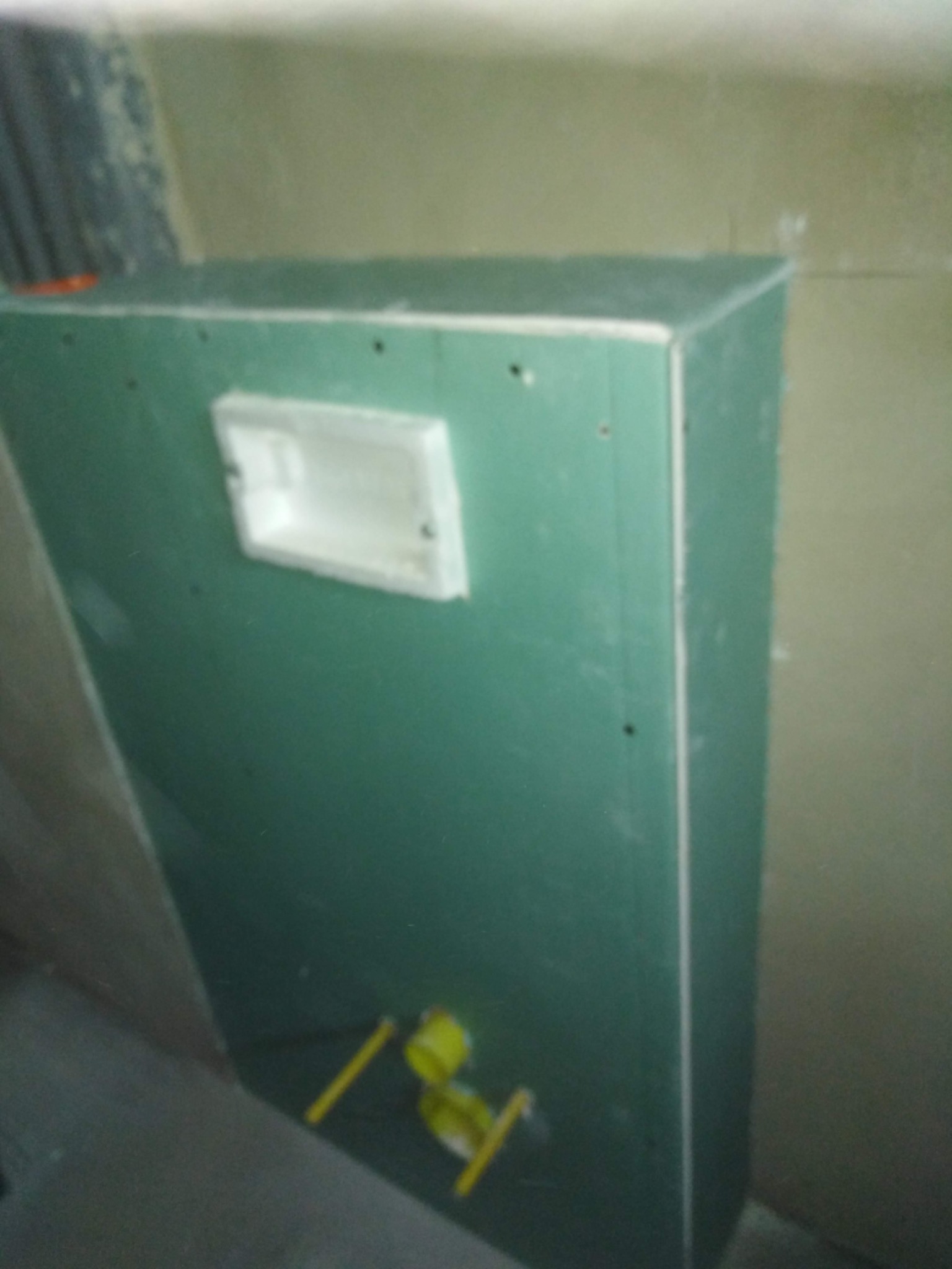 Zabudowa z zielonej płyty gipsowo-kartonowej, z widocznym otworem rewizyjnym i żółtymi rurami instalacji wodnej, w trakcie prac wykończeniowych.