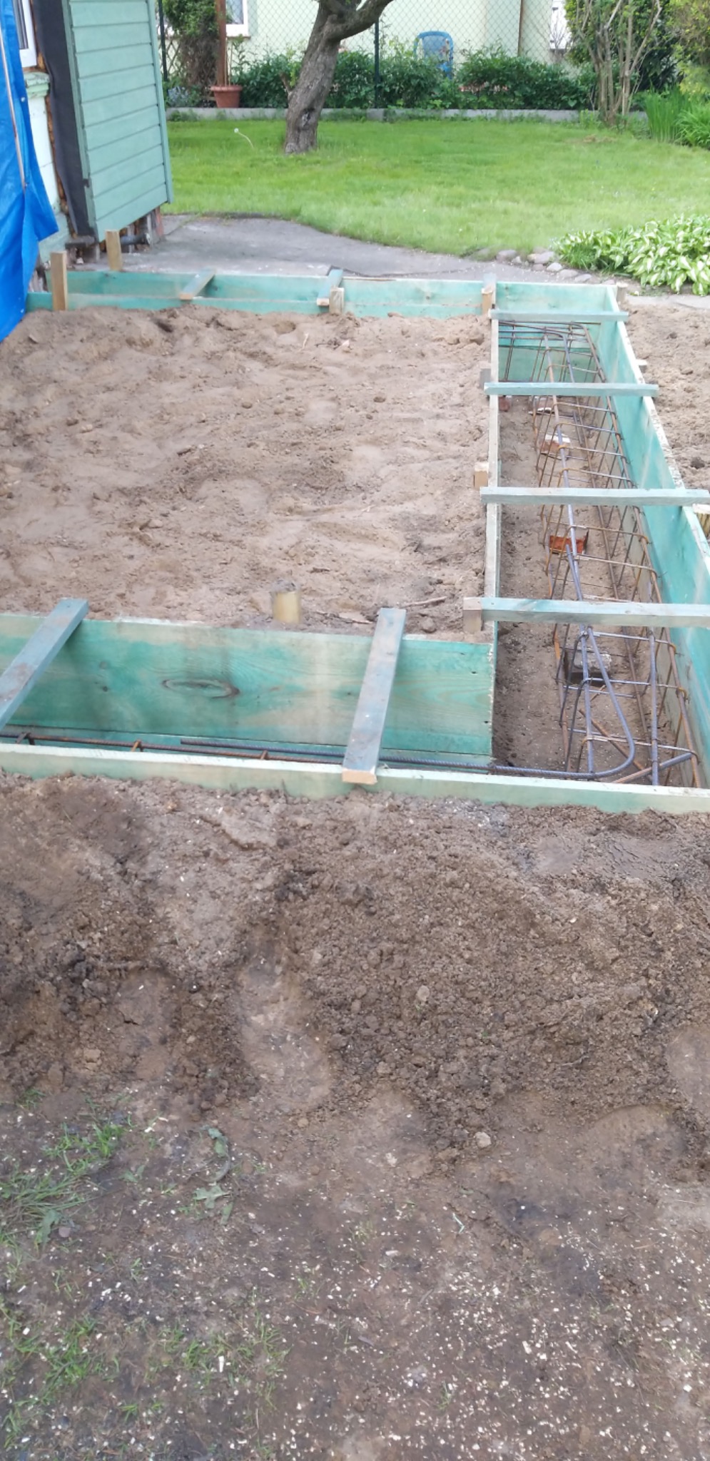 Przygotowanie fundamentów: szalunek z desek pomalowanych na zielono, wypełniony zbrojeniem stalowym, częściowo zasypany piaskiem na trawniku przed domem.