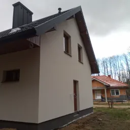 Świeżo otynkowany, dwukondygnacyjny dom z ciemną stolarką okienną i czarnym kominem, widok z boku na tle drugiego, budowanego domu z czerwoną dachówką.