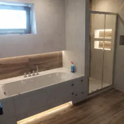 Nowoczesna łazienka z wanną obudowaną płytkami, drewnopodobną podłogą, kabiną prysznicową i subtelnym oświetleniem LED pod wanną. Na blacie obok wanny stoi niebieski płyn do czyszczenia.