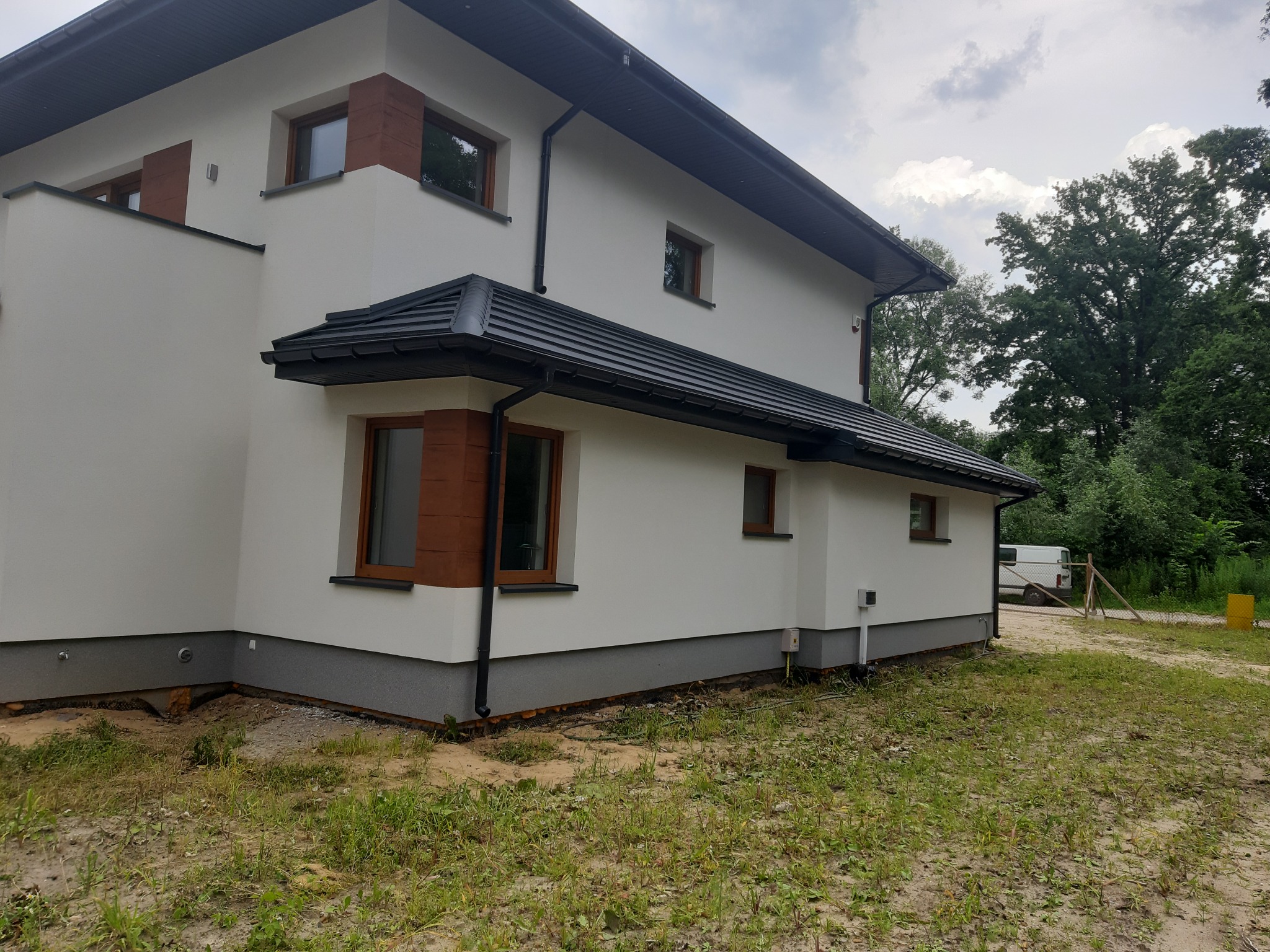 Nowoczesny, dwukondygnacyjny dom z jasną elewacją, ciemną dachówką i drewnianymi akcentami okiennymi, widoczny z boku na tle zieleni.