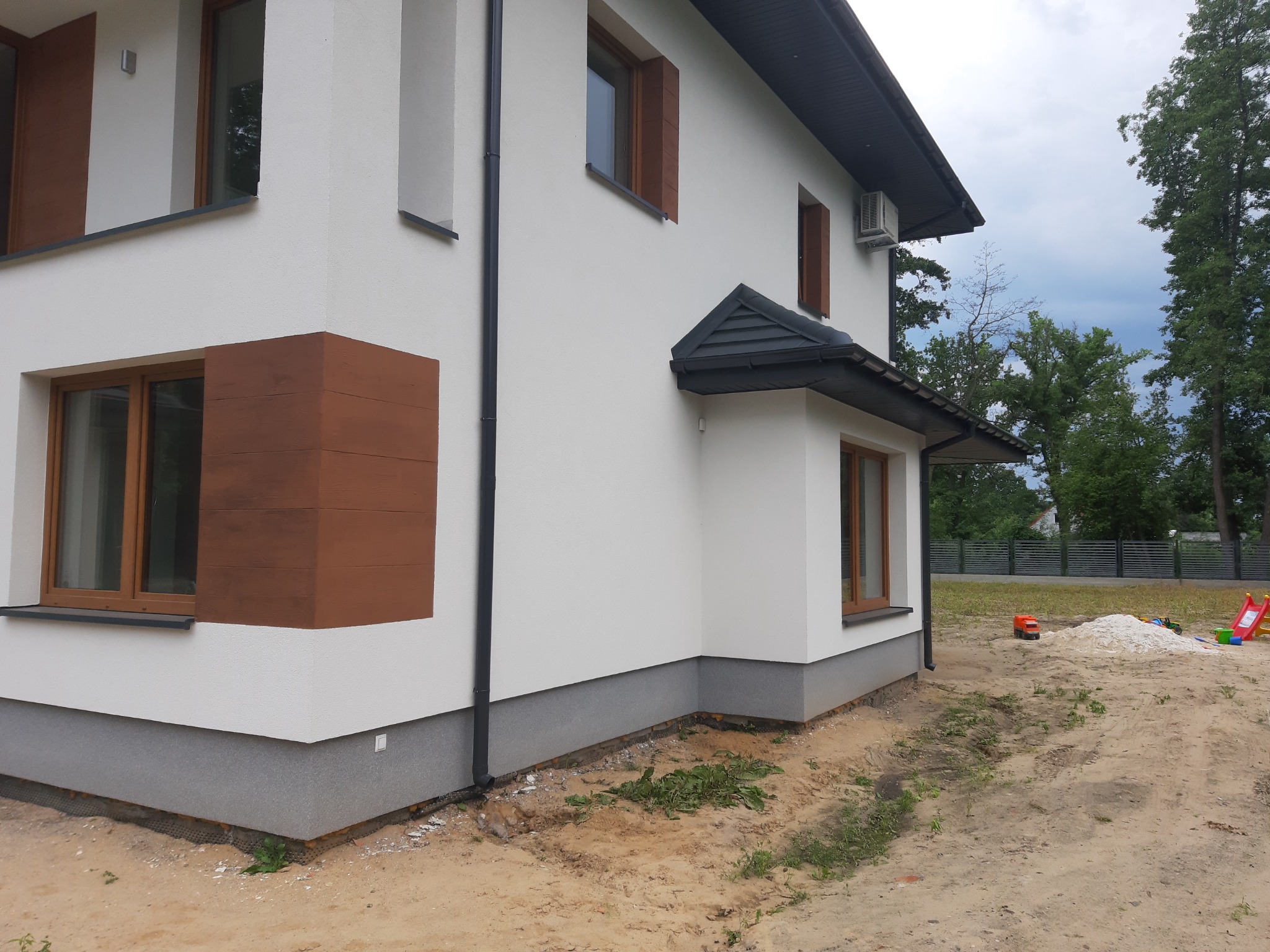 Nowoczesny dom jednorodzinny z białą elewacją, drewnianymi elementami okiennymi i szarą podmurówką. Ujęcie z zewnątrz, widoczny fragment ogrodu z piaskiem i zabawkami.