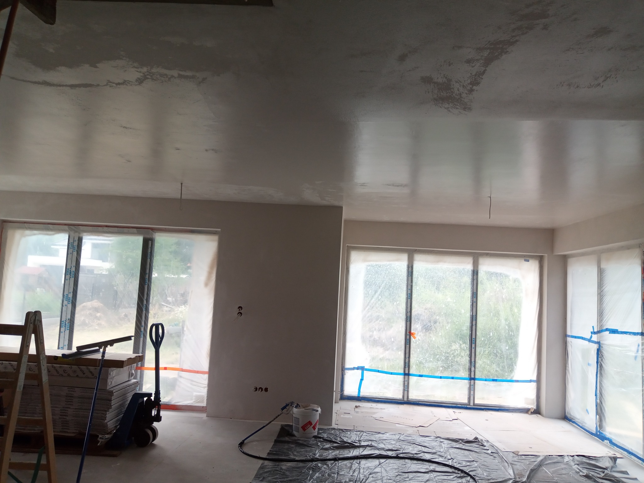 Wnętrze w trakcie tynkowania maszynowego: świeżo otynkowany sufit i ściany, okna zabezpieczone folią, widoczny agregat tynkarski, drabina i paleta płyt gipsowo-kartonowych.