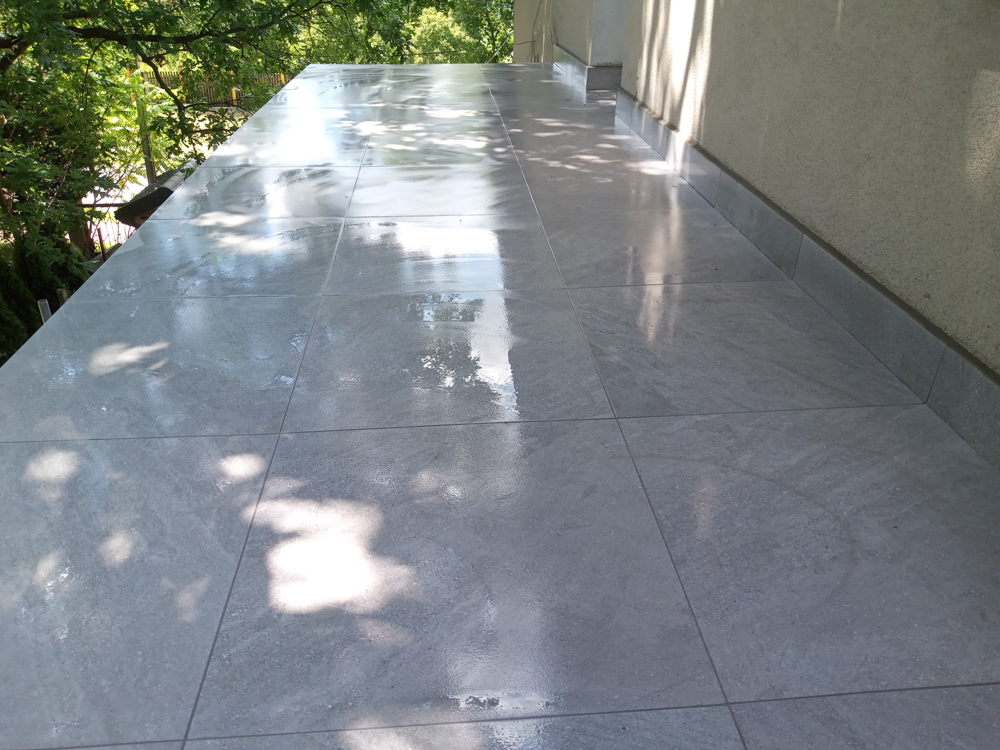 Wyłożony szarymi płytkami taras z widokiem na zieleń drzew, z refleksami światła słonecznego na powierzchni płytek.