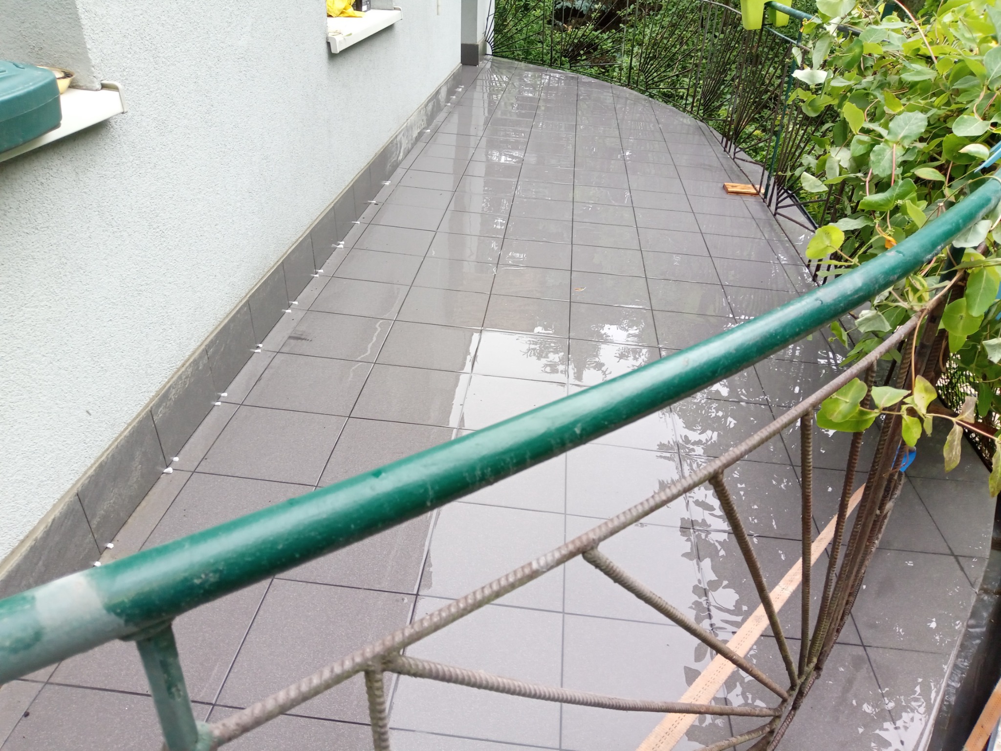 Wykładanie szarych płytek na mokrym, zakrzywionym balkonie z zieloną metalową barierką i widokiem na zieleń.