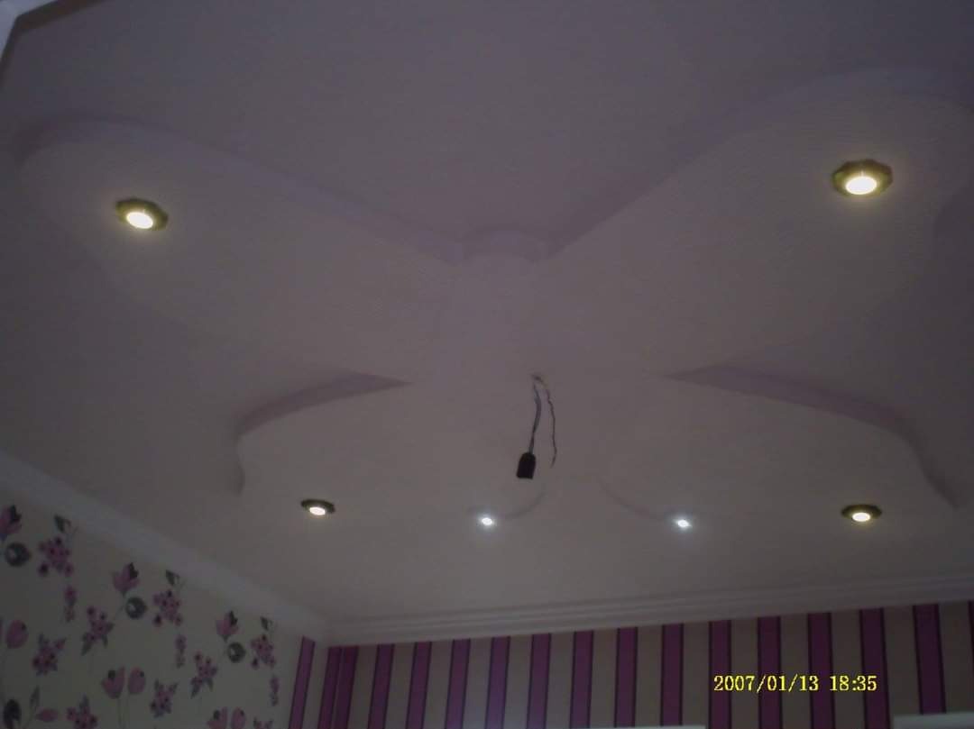 Ozdobny sufit z nieregularnymi kształtami i punktowym oświetleniem, zwisający przewód w centralnej części, ściany w kwiaty i pionowe pasy.