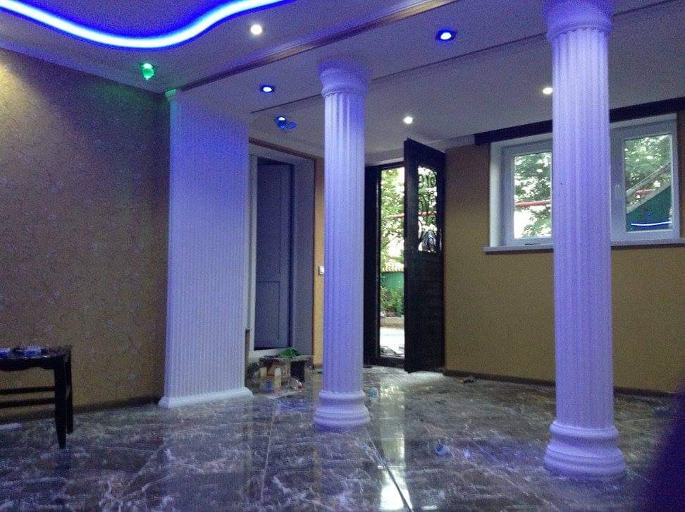 Wnętrze pokoju z marmurową podłogą, dwiema kolumnami w stylu greckim, podświetlanym sufitem LED, otwartymi drzwiami balkonowymi i oknem.