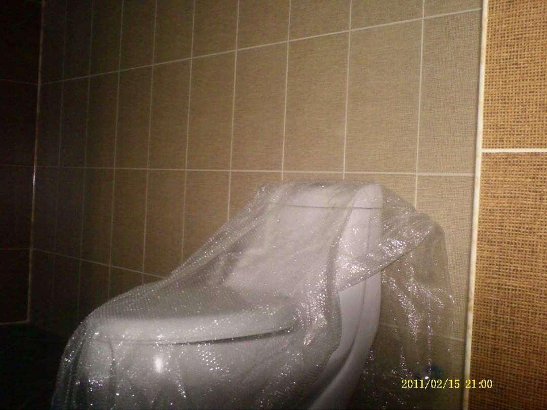 Nowa, biała toaleta zabezpieczona folią ochronną, na tle wykafelkowanej ściany z brązowych płytek z jasną fugą i dekoracyjnej ściany z teksturą lnu.