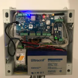 Otwarta skrzynka z elektroniką systemu alarmowego: płyta główna z modułem GSM, moduł rozszerzeń, zasilacz impulsowy i akumulator podtrzymujący, widoczne diody LED sygnalizujące pracę.