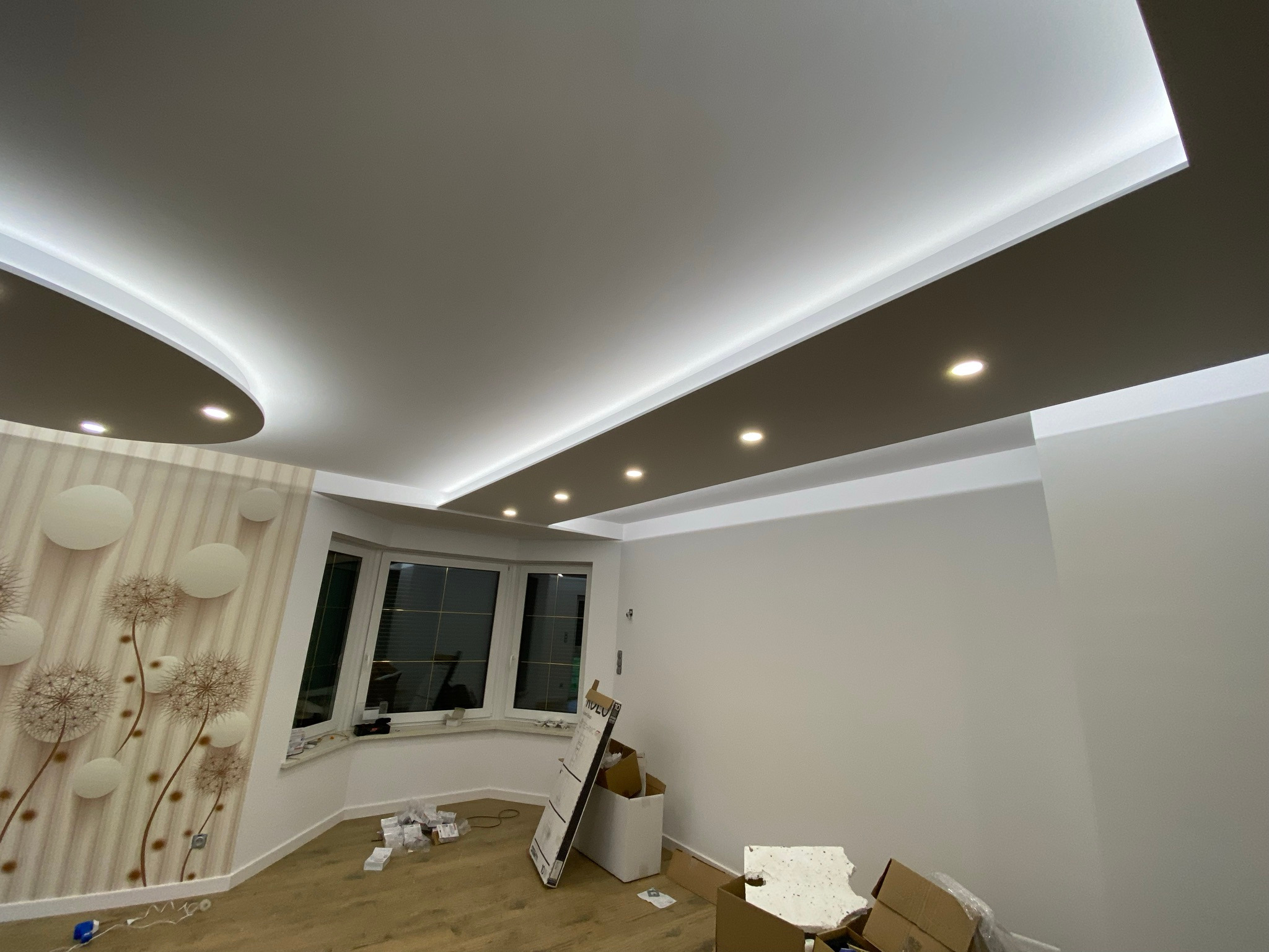 Nowoczesny sufit podwieszany z oświetleniem LED, widok na okno wykuszowe i fragment tapety z motywem dmuchawców w jasnym pokoju, w trakcie prac wykończeniowych.