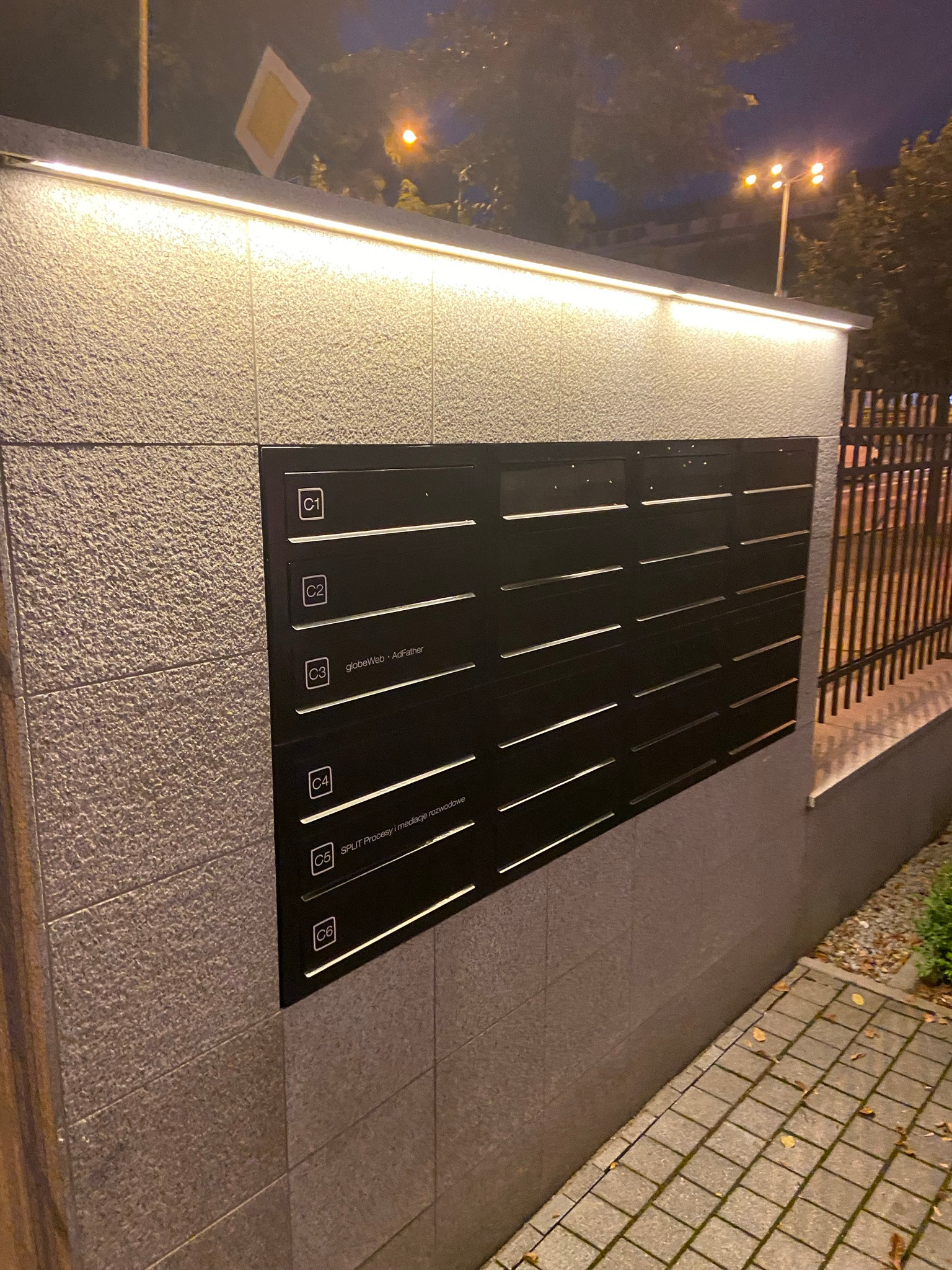 Zestaw czarnych skrzynek pocztowych na elewacji budynku, oświetlonych listwą LED, z widocznymi nazwami firm na niektórych skrzynkach, w tle fragment chodnika i ogrodzenia.